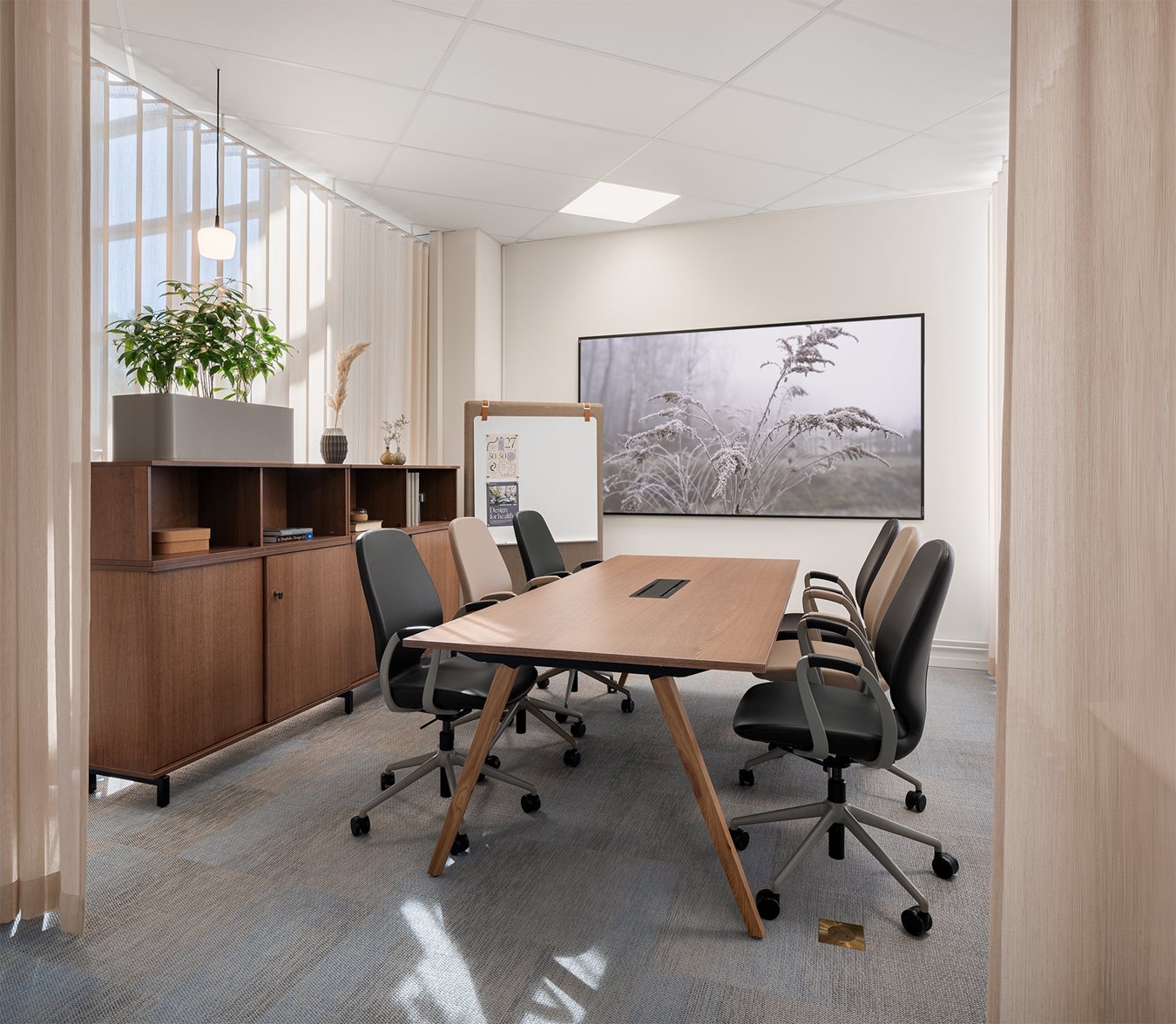 Ett modernt konferensrum med ett rektangulärt mötesbord i trä, svarta och beige kontorsstolar med hjul, en stor mötesrumskärm på väggen och en förvaringsmöbel med växter. Rummet har ljusa väggar, en textil matta och mjuk belysning.