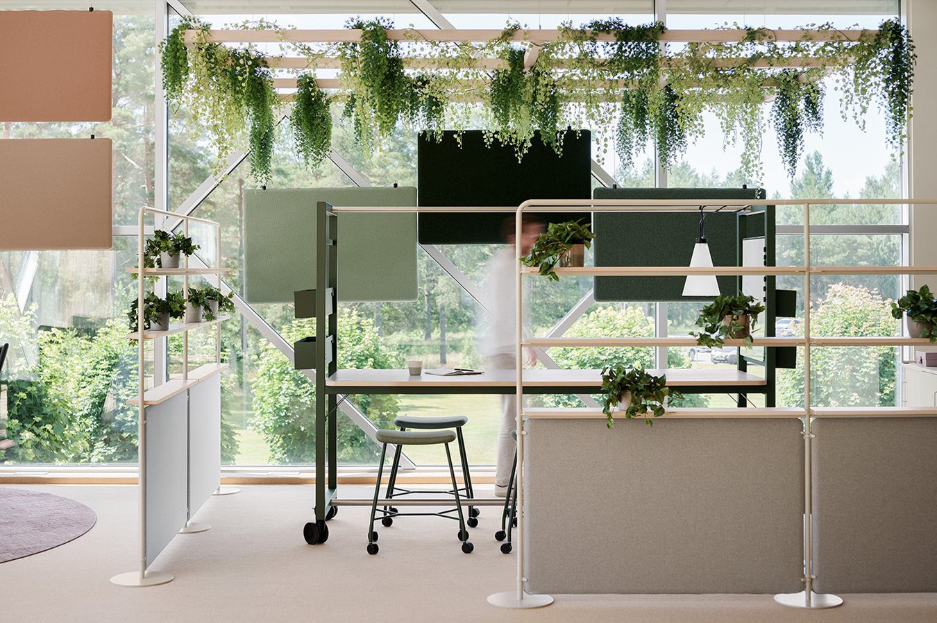 En öppen och flexibel projektplats med stort mötesbord, ljusa takabsorbenter, golvskärmar och grönskande växter som hänger från taket. Stora fönster släpper in naturligt ljus och ger utsikt mot grönska utanför.