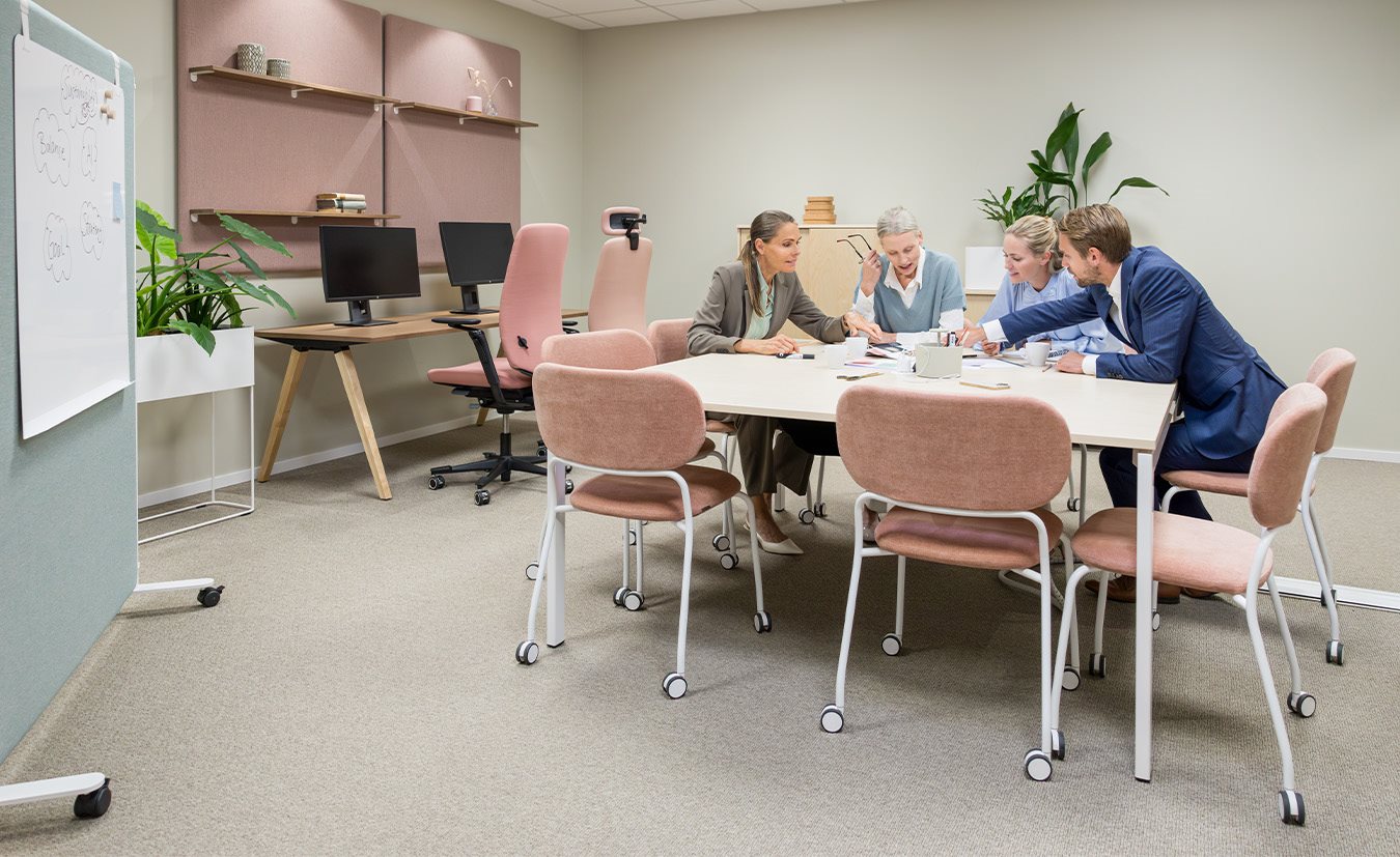 Ett mötesrum med ett stort vitt mötesbord omgiven av rosa och beige stoppade stolar med hjul. Fyra personer har en diskussion vid bordet. I bakgrunden finns skrivbord med datorer, växter och ljudabsorbenter på väggen.